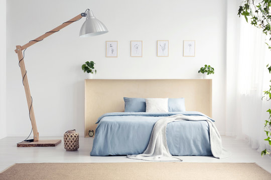 Designer lamp next to blue bed with blanket in white bedroom interior with posters. Real photo