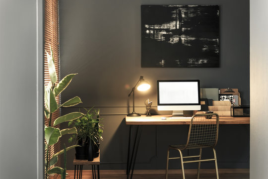 Gold chair standing by the wooden desk with lamp, notebooks and mockup computer in real photo of dark living room interior with modern poster on the wall