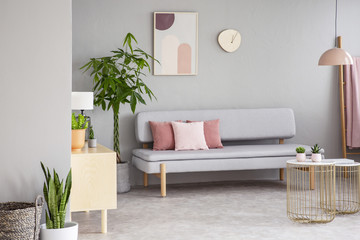 Real photo of grey living room interior with lounge with pink cushions, gold end tables, fresh...