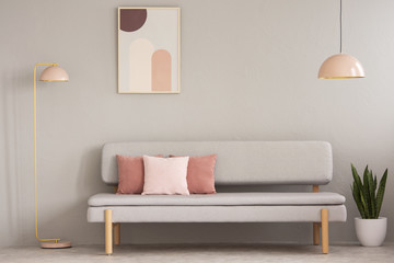 Real photo of grey sitting room interior with couch with cushions, pastel pink lamps, fresh plant and simple poster on the wall