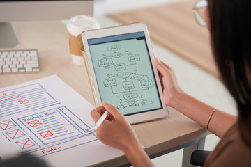 Fototapeta premium Crop back view of woman sitting at desk with website layout and looking at program flowchart diagram on tablet screen in hands