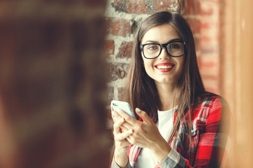 Cute teenager long-hair female girl in glasses rstaying with mobile phone and smiling near the window in the loft modern office