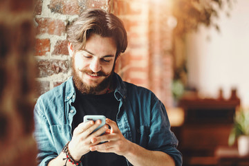 Handsome young bearded boy reading sms on the mobile phone and smiling near the window in the loft modern office