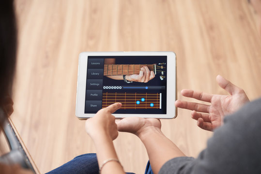 Crop back view of man and woman holding tablet with video of playing guitar tutorial and discussing it pointing at screen 