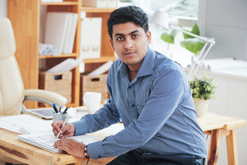 Indian ethnic office employee sitting at desk and looking at camera in office while doing paperwork