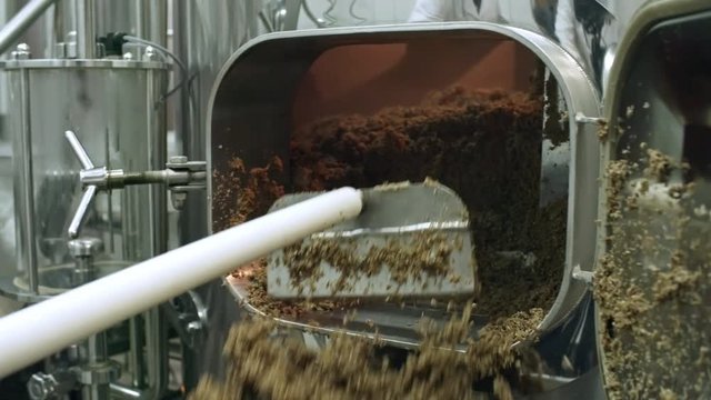 Tracking closeup shot of hands of brewery worker removing grist from milling machine with metal shovel