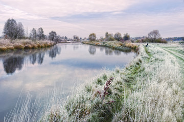 Beautiful Autumn Belarusian Landscape On The Topic Of Fishing: Lone Fisherman Sits On The Grassy Bank Of The River, Covered With Hoarfrost. On Fishing: Waiting For A Bite. Cold Autumn Morning, Belarus