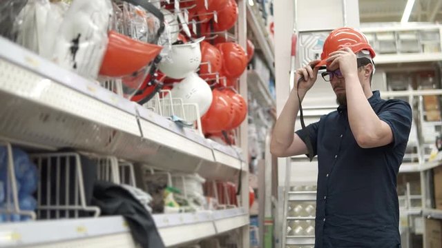 Bearded guy is choosing construction helmet in hypermarket for builders. He is standing in a hall in shop and trying on orange hat