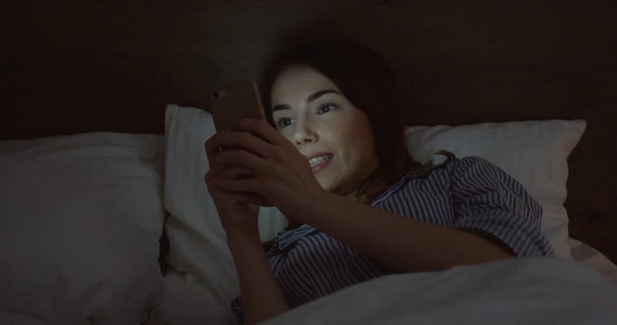 Close up of the charming Caucasian woman with dark hair lying in the bed, chatting on the smartphone late in the night and laughing. Inside