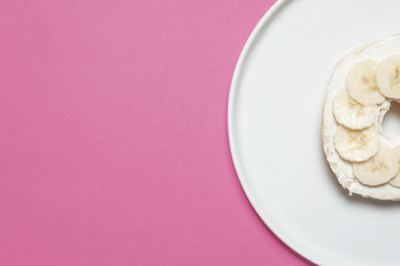 Half a banana and cream cheese bagel on a white plate on a pastel pink background
