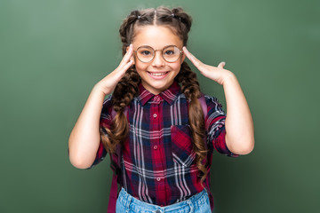 smiling schoolchild touching head and looking at camera near blackboard