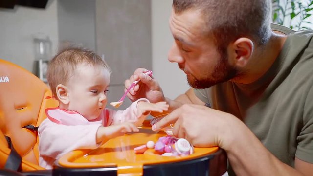 Handsome young father feeding to his baby food in the kitchen. He is making faces and feeding his daughter. Father and daughter time.