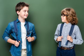 schoolboys with backpacks looking at each other near blackboard