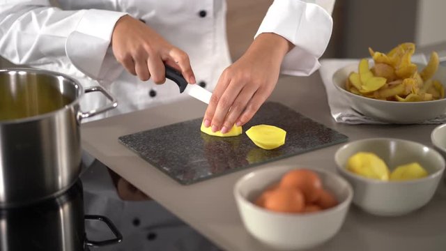 4K cooking footage, close up woman in cooks jacket female hands cutting raw peeled potatoes on cutting board with ceramic knife
