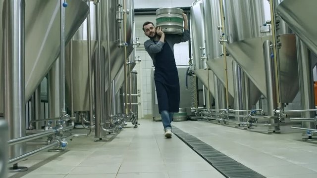 Tracking shot of male worker carrying beer keg on shoulder and walking through brewery towards the camera