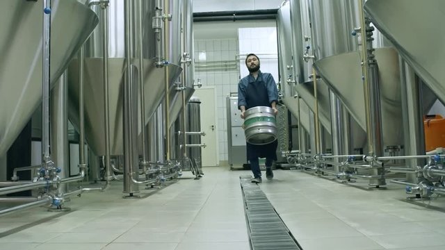 Bearded Asian man in uniform carrying beer keg while walking through brewery towards the camera