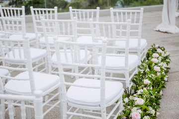 Close-up back side of white chiavari chairs arrangement for modern wedding ceremony, white and pink roses and green leaf decoration on the aisle walk way