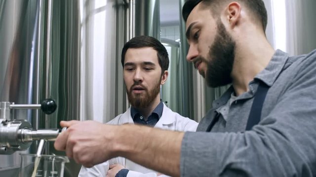 Tilt down of worker pouring freshly brewed beer from vessel into jug in brewery while his colleague in lab coat explaining something