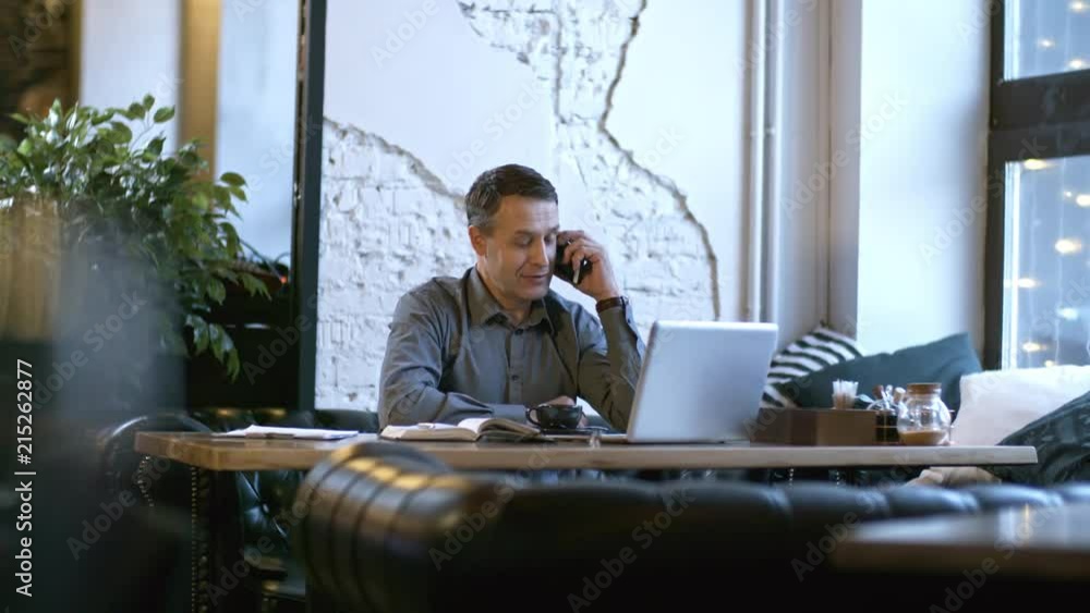 Poster tracking shot of smiling middle-aged man enjoying refreshing coffee at table in cafe and talking on 