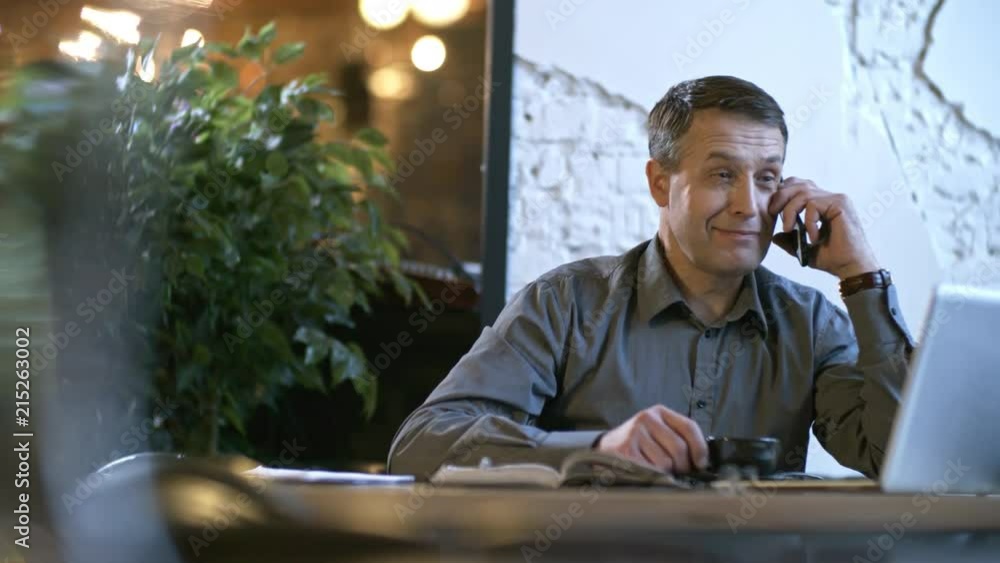 Poster Tracking shot of middle-aged man having telephone conversation and smiling when sitting at table in cafe