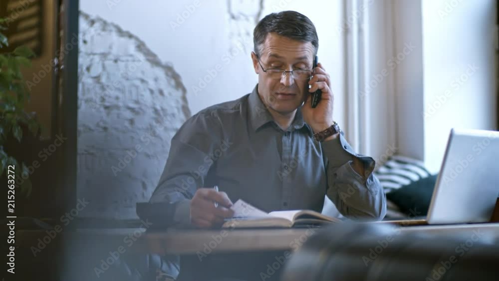 Poster tracking shot of serious middle-aged businessman in glasses taking notes in notebook when talking on