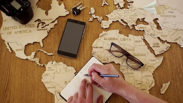 A close portrait of a man writes a plan for his journey. Wooden world map layout on the table, notebook, pen, phone, keys, glasses and camera