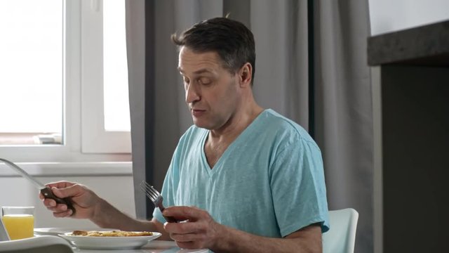 Medium shot of middle-aged man sitting at table in kitchen and talking to someone before eating scrambled eggs for breakfast