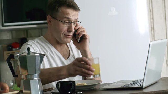 Handheld shot of multi tasking male freelancer talking on the phone and using laptop computer while having breakfast at home