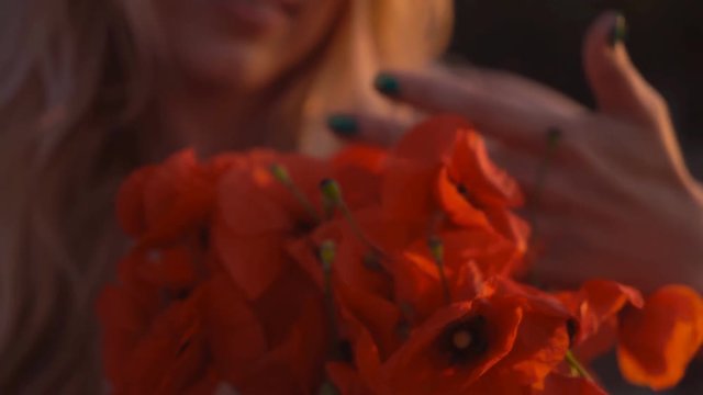 cute blonde girl in red dress is standing at sunset. A woman is holding a bouquet of red poppies. close-up portrait. Person stands on the bridge. 4K
