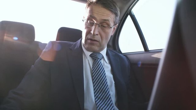 Medium shot of busy mature man in glasses and business suit using laptop computer and smartphone when going to work by taxi