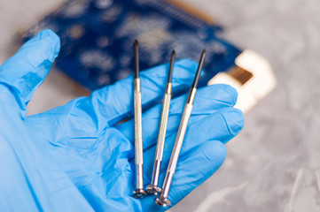 Mens hand in blue rubber glove holds three small metal screwdrivers on background of pc graphics card with copper radiator on old gray concrete floor