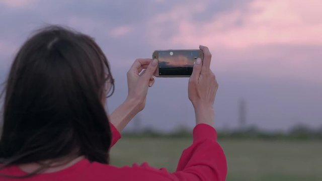 Beautiful woman takes photos of sunset on field, using smartphone, urning towards the camera. Close-up. In windy weather.