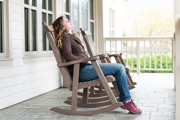 Young woman sitting on reclining, rocking chair, recliner, rocker by house, home, townhouse with closed eyes, enjoying fresh air, nature, sunset outside, outdoors