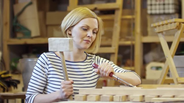 Tracking medium shot of blonde woman hitting chisel with mallet to shape wood in carpentry workshop