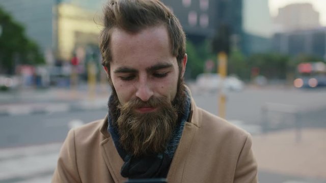 close up portrait of attractive stylish young bearded hipster man texting browsing using smartphone messaging app in city street