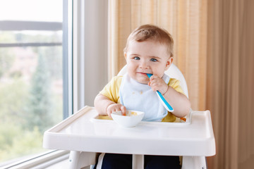 Portrait of cute adorable Caucasian child boy with dirty messy face sitting in high chair eating apple puree with spoon. Everyday home childhood lifestyle. Infant trying supplementary baby food