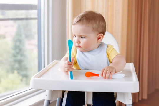 Portrait of cute adorable Caucasian child boy sitting in high chair with spoons. Everyday home childhood lifestyle. Infant trying supplementary baby food