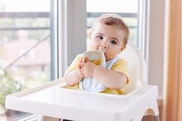 Portrait of cute adorable Caucasian child boy with dirty messy face sitting in high chair eating apple puree with fingers. Everyday home childhood lifestyle. Infant trying supplementary baby food