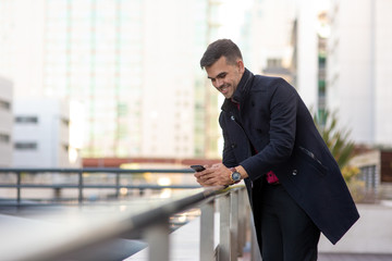 Cheerful handsome Hispanic man smsing on smartphone and leaning on railing in city. Positive...