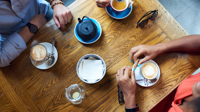 Overhead of unrecognizable people hanging out in cafe with cups of coffees on wooden table.