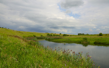 Obraz premium Cloudy summer landscape with fields,meadows,woods and small river.Dark stormy clouds in dramatic overcast sky.Rainy season.