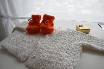 Gifts for new born baby in winter: a pair of  orange wool shoes, a handmade woven white pullover and a golden gift box.