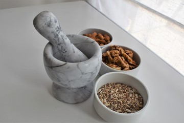 Three white containers with almonds, walnuts and a mixture of sunflower, sesame and chia seeds near a marble mortar.