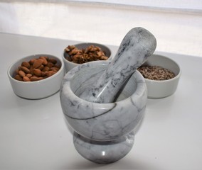 Three white containers with almonds, walnuts and a mixture of sunflower, sesame and chia seeds near a marble mortar.