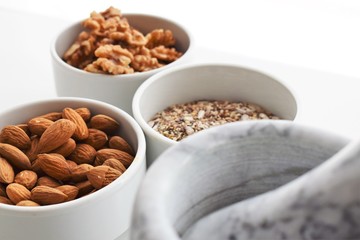 Three white containers with almonds, walnuts and a mixture of sunflower, sesame and chia seeds near a marble mortar.