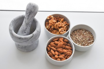 Three white containers with almonds, walnuts and a mixture of sunflower, sesame and chia seeds near a marble mortar.