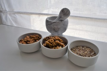 Three white containers with almonds, walnuts and a mixture of sunflower, sesame and chia seeds near a marble mortar.