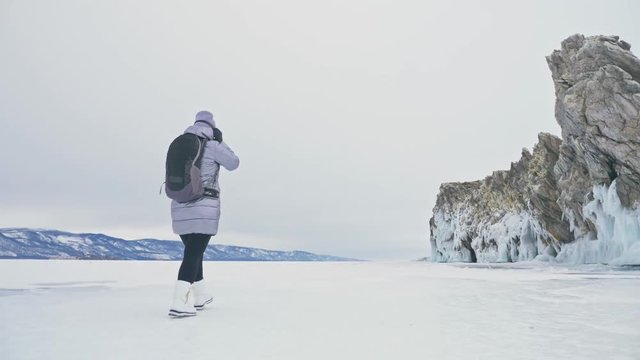 Travel of woman on ice of Lake Baikal. Trip to winter island. Girl is walking at foot of ice rocks. Traveler looks at beautiful ice grotto. Hiker wears sports glasses, silver jacket, backpack. Extreme