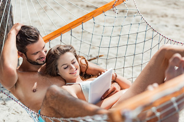 smiling couple using digital tablet while relaxing on hammock on beach