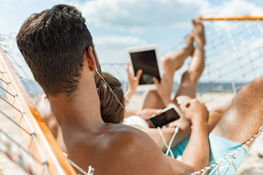 young couple listening music with earphones and gadgets while relaxing on hammock on beach